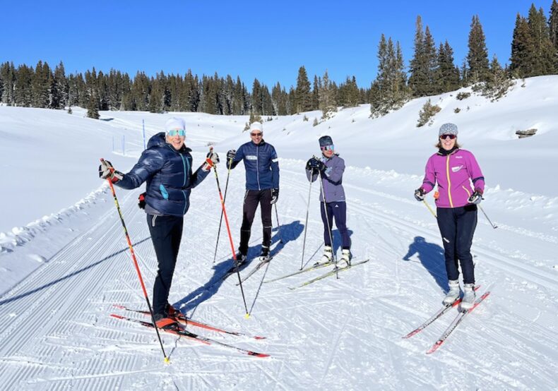 
Photograph of four people, including Christie, pausing mid-ski to smile on a beautiful snowy day.