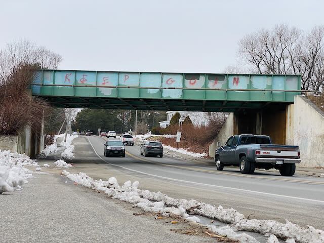 Photo of a battered green overpass above a 2-lane road with cars in either direction. The sky is gray-white and the trees are bare. There is snow on the ground. Red letters spray-painted on the overpass read "KEEP GOIN"