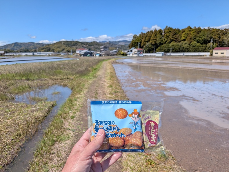 Hand holding a bag of snacks; trail, flooded rice fields, mountains, and trees in the background. 